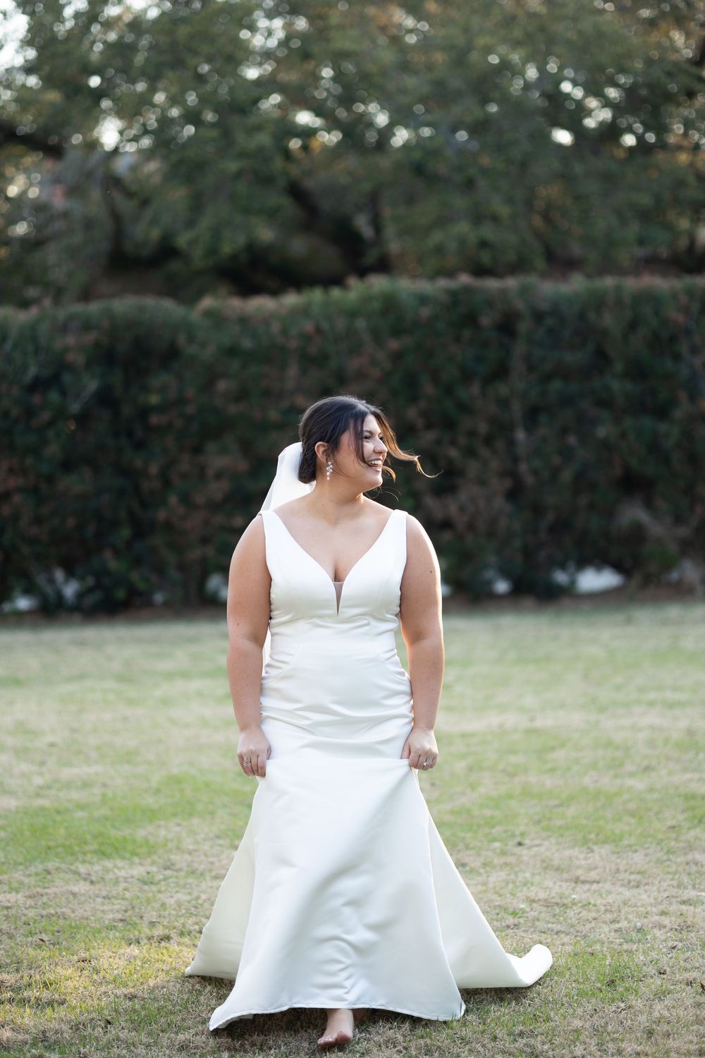 Bride in white mermaid wedding gown with veil smiles outdoors on green lawn with hedge backdrop.