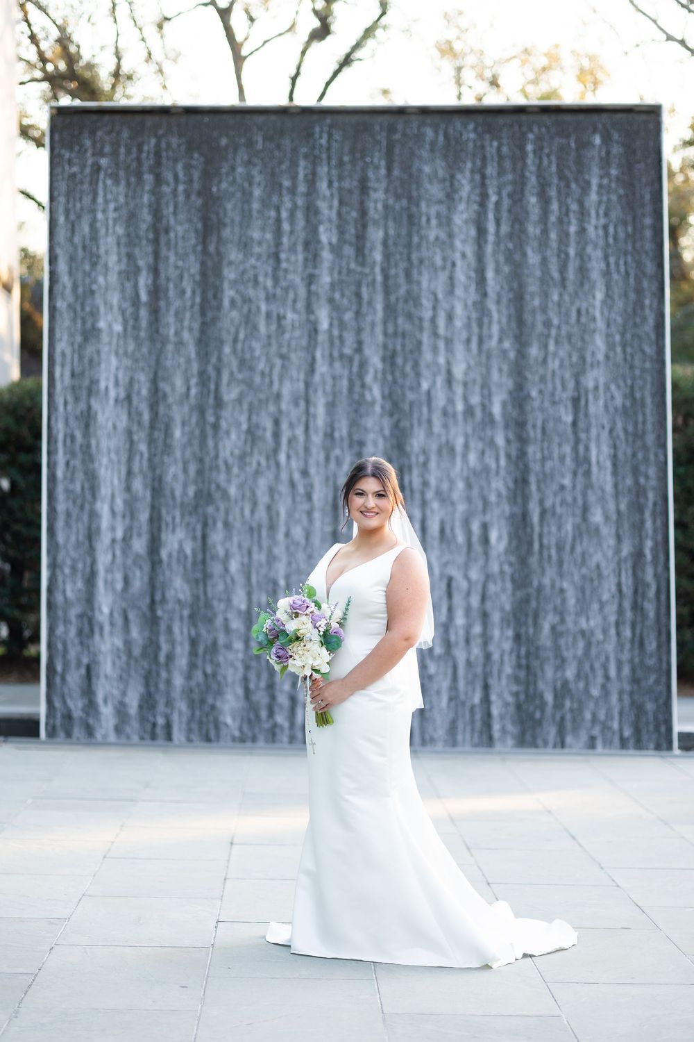 Bride in white gown holding purple and white bouquet stands before a large water wall fountain outdoors.
