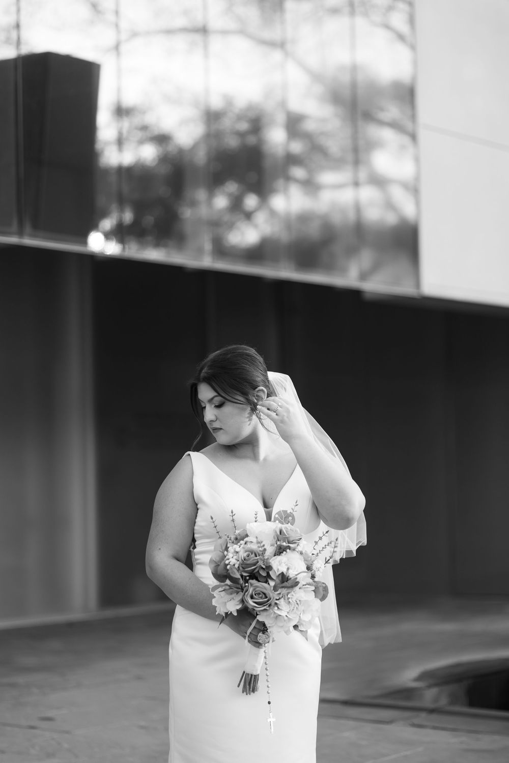 Black and white photo of a bride in a white gown holding a bouquet, gently touching her hair outdoors.