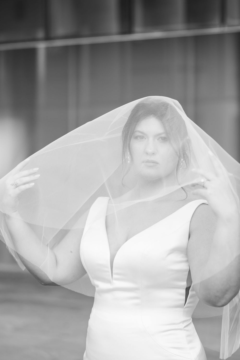 Black and white portrait of a bride in a white gown lifting her veil, gazing directly at the camera.