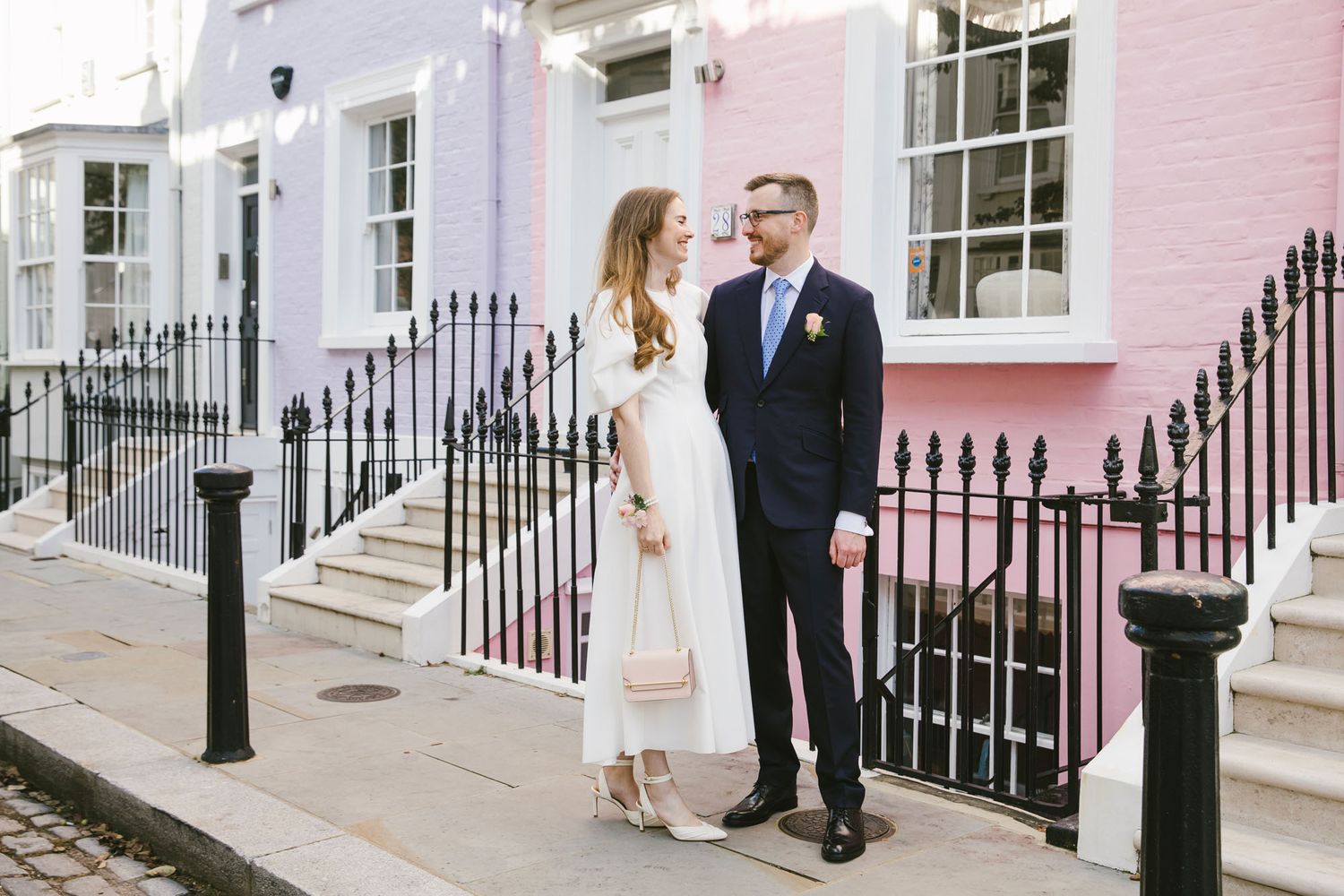 Elegant couple posing in front of colorful pastel townhouses on a charming London street.