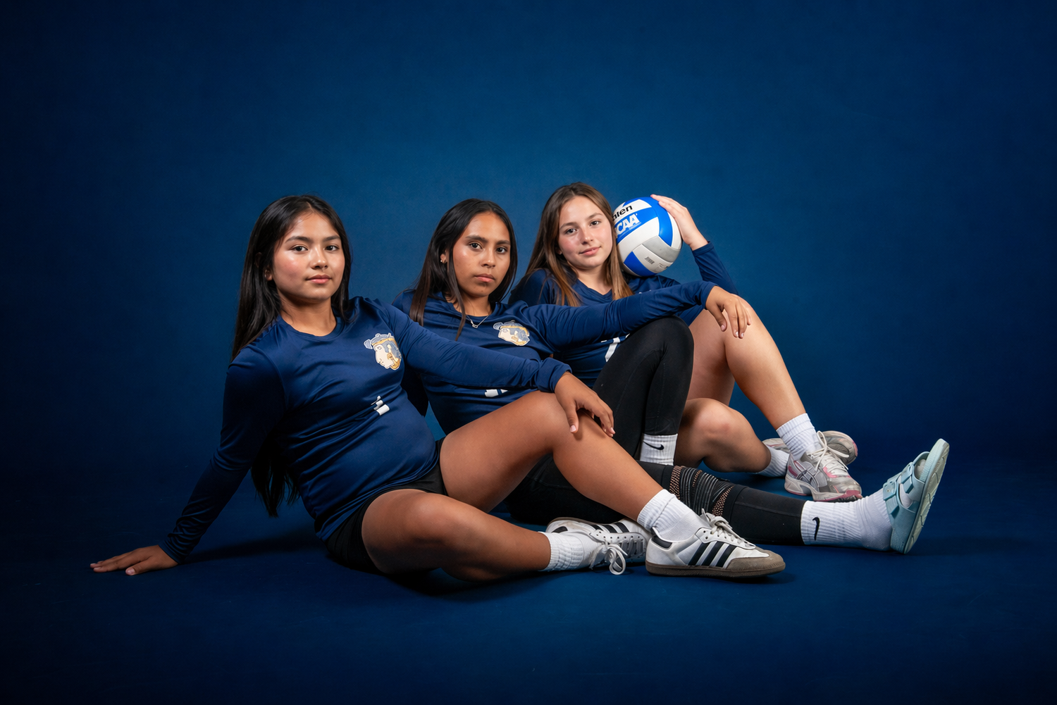 Three female volleyball players in navy uniforms pose together on a blue studio background, one holding a volleyball.