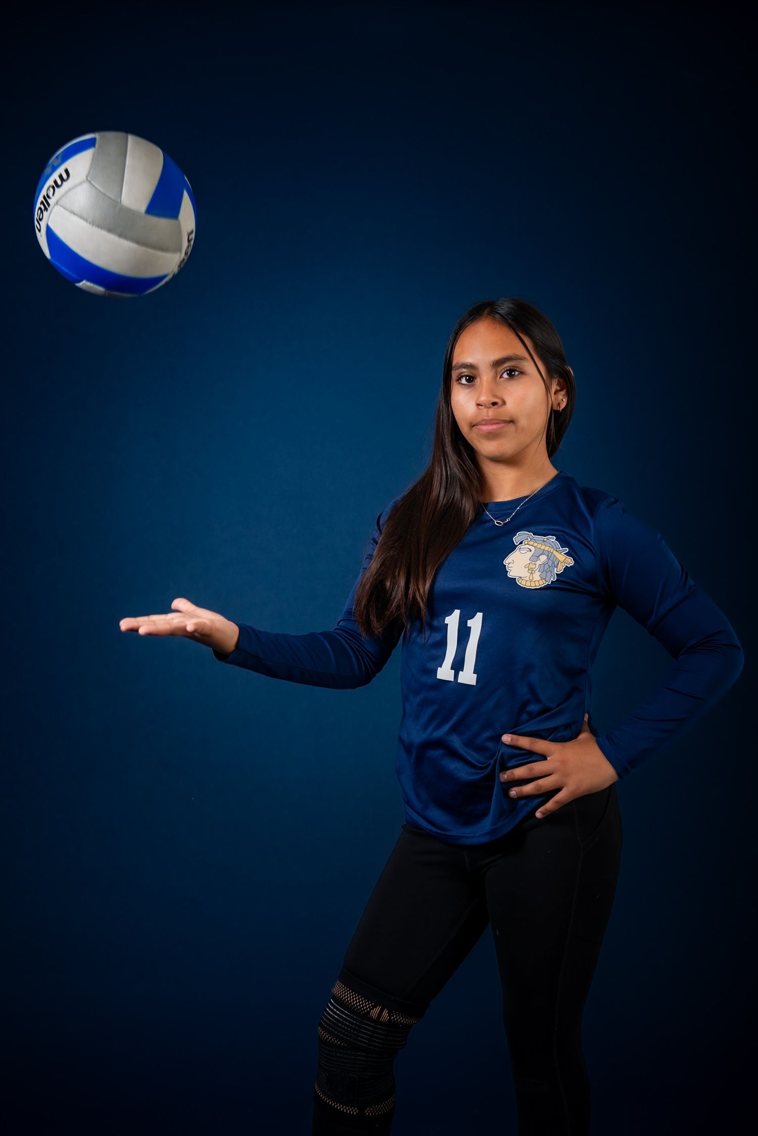 Female volleyball player #11 in blue jersey poses with a volleyball against a dark blue background.