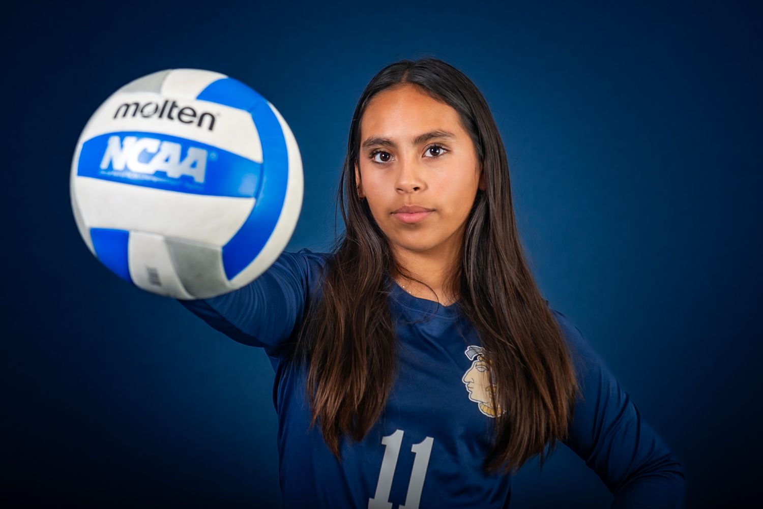 Female volleyball player in blue jersey number 11 holding a Molten NCAA volleyball against a blue background.