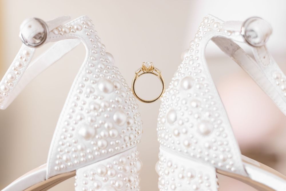 White pearl-embellished wedding heels with a gold diamond engagement ring nestled between them on a soft background.