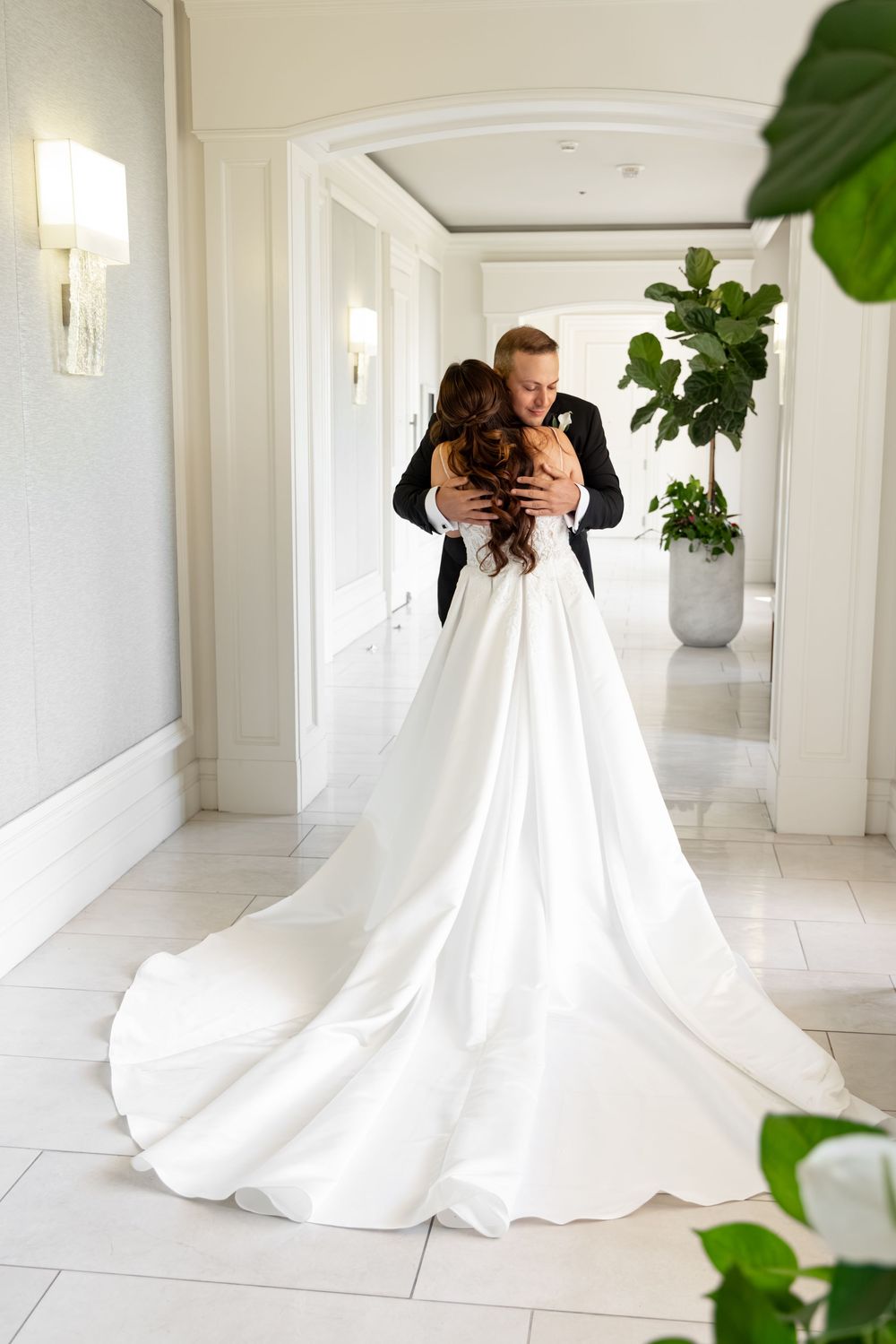 Bride in elegant white gown with train embraces groom in black suit in a bright, luxurious hotel hallway.