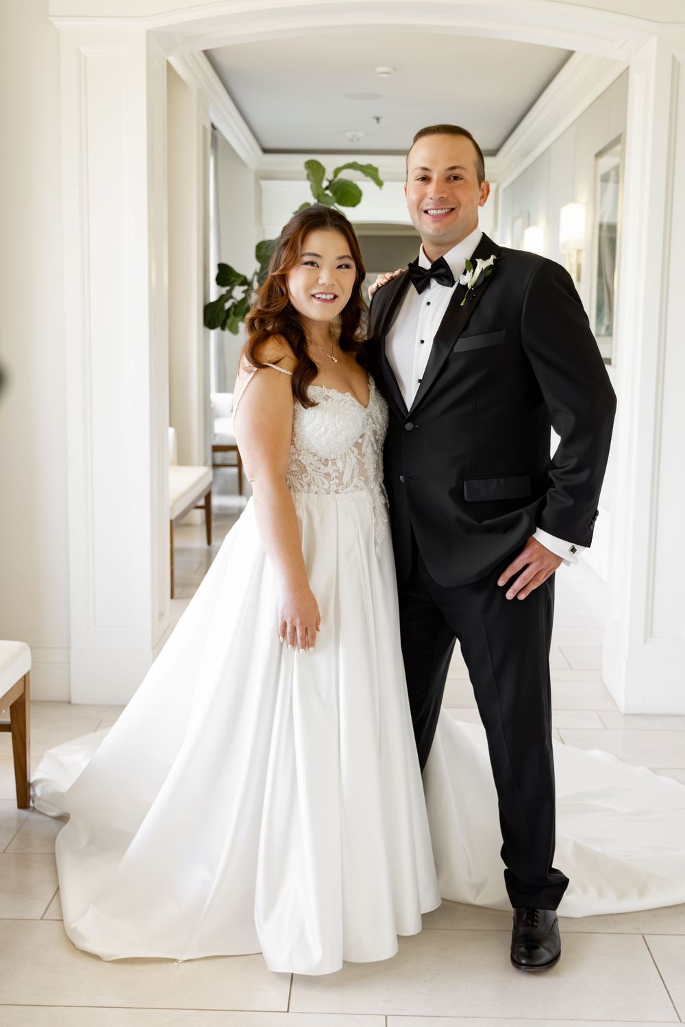 Smiling bride in beaded white gown poses with tuxedo-clad groom in a bright elegant venue hallway.