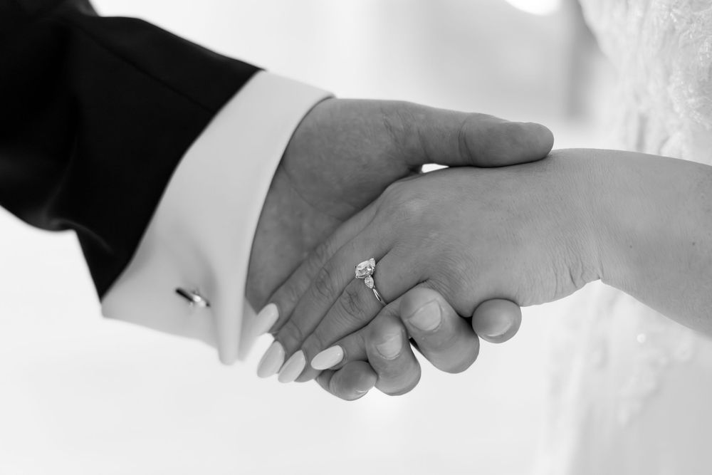 Black and white photo of bride and groom holding hands, showcasing a diamond engagement ring on wedding day.