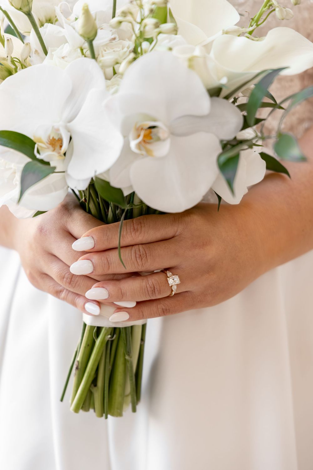 Bride holding white orchid wedding bouquet with diamond engagement ring and white manicured nails.