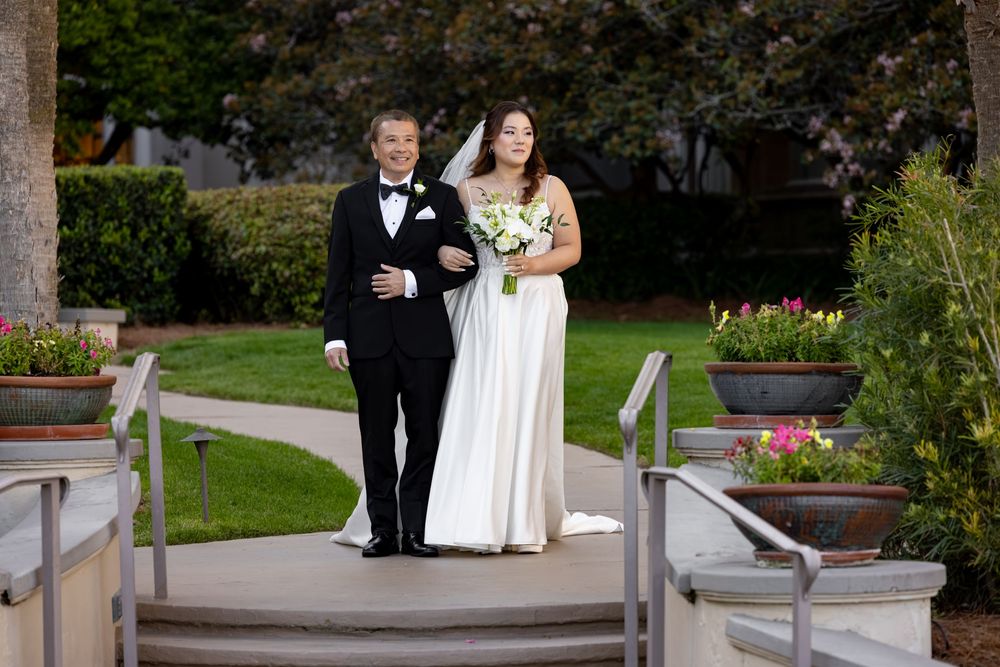 Bride in white gown holding bouquet walks with groom in tuxedo at outdoor wedding ceremony.