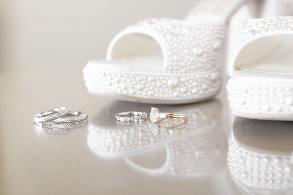 White pearl-embellished bridal heels displayed with diamond wedding rings and gold bands on a reflective surface.