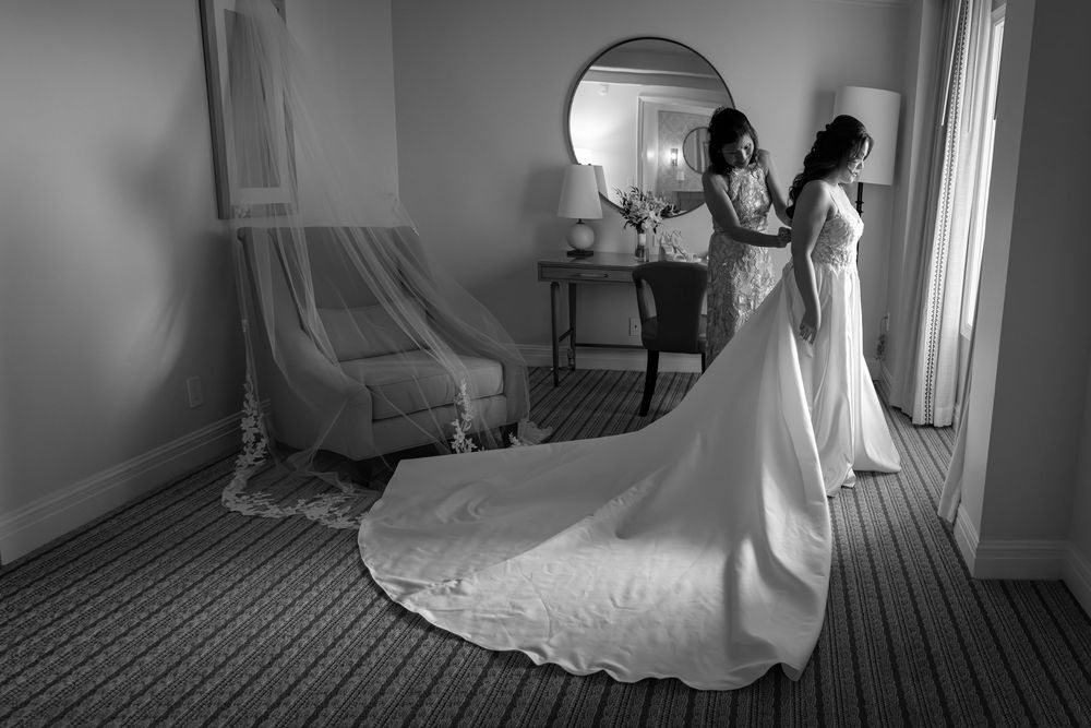 Bride in elegant gown with long train being helped by bridesmaid in black and white hotel room.