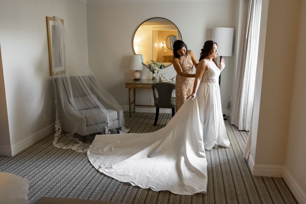 Bride in a white gown with long train being helped by bridesmaid in elegant hotel room with mirror.