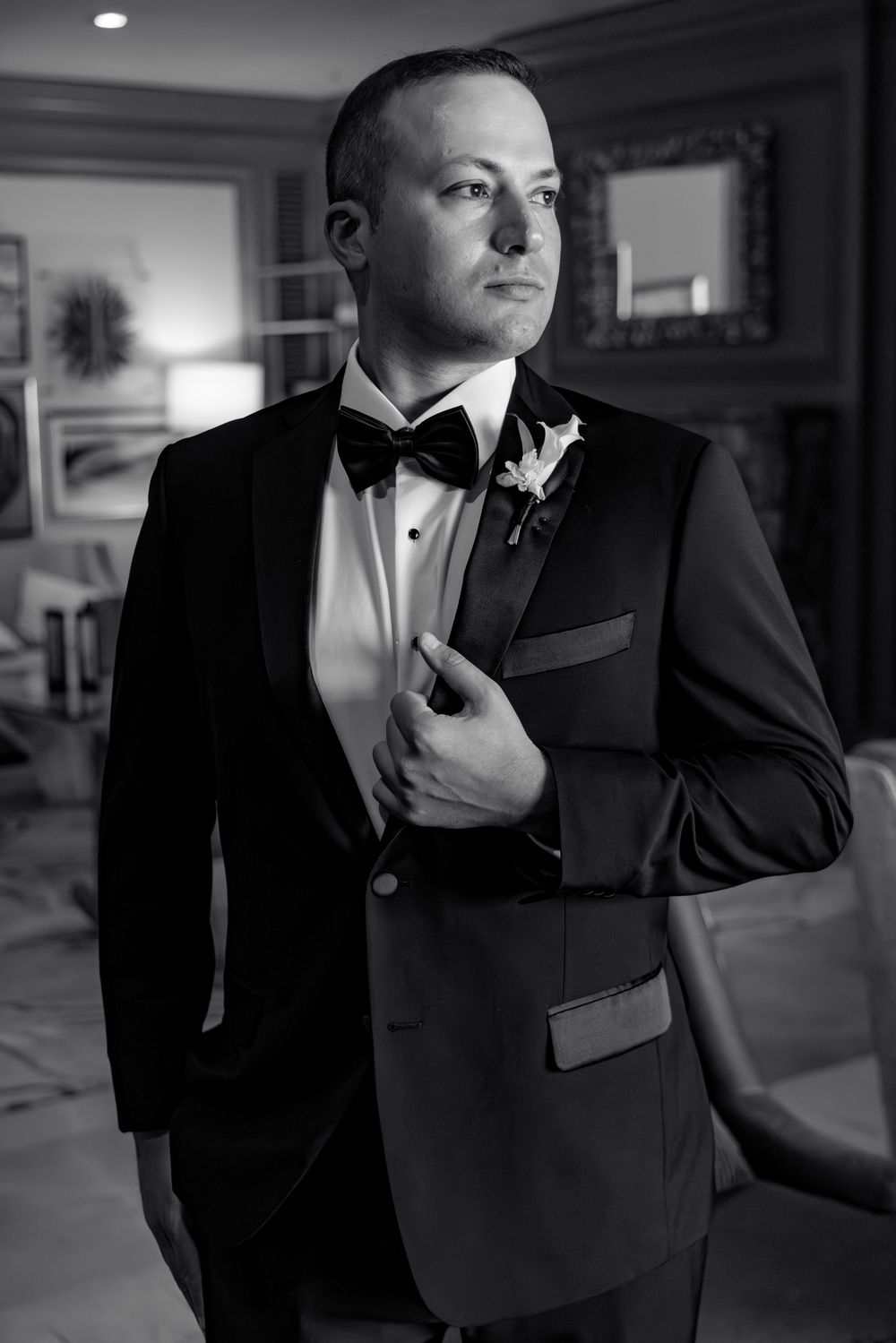 Groom in black tuxedo with bow tie and boutonniere adjusting jacket in elegant room.