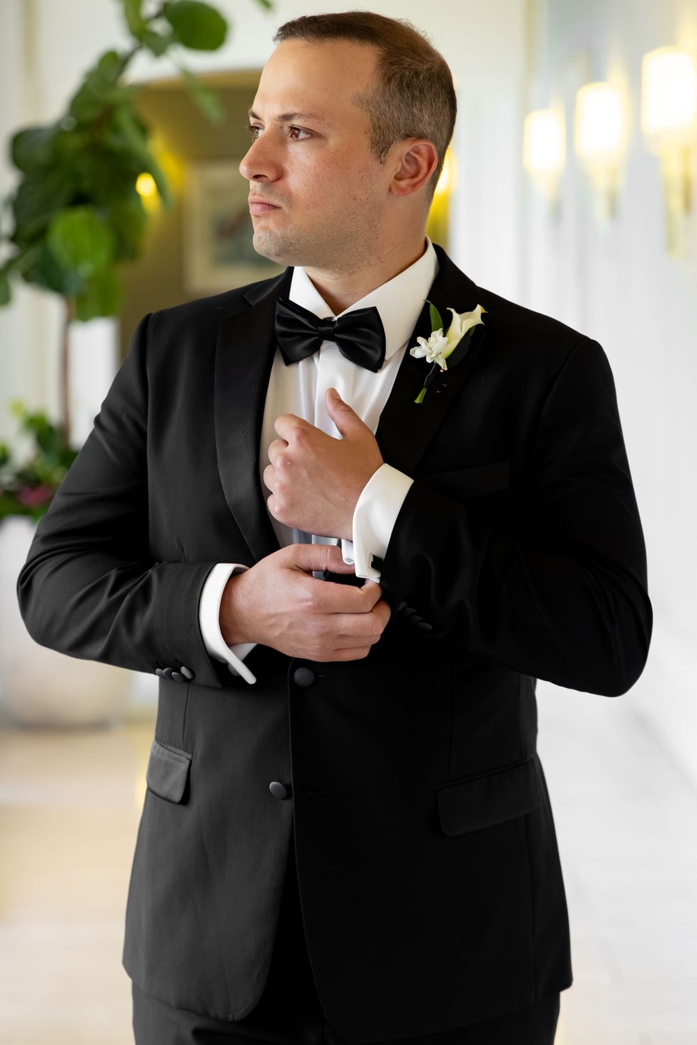 Dapper groom in black tuxedo with bow tie and white boutonniere adjusting cufflinks indoors.