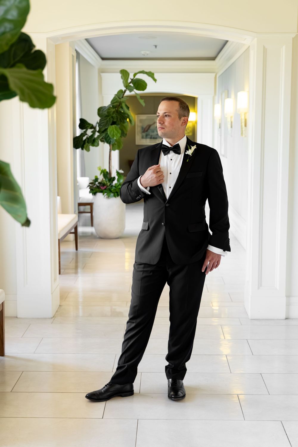 Groom in black tuxedo with bow tie and boutonniere standing in elegant hotel corridor.