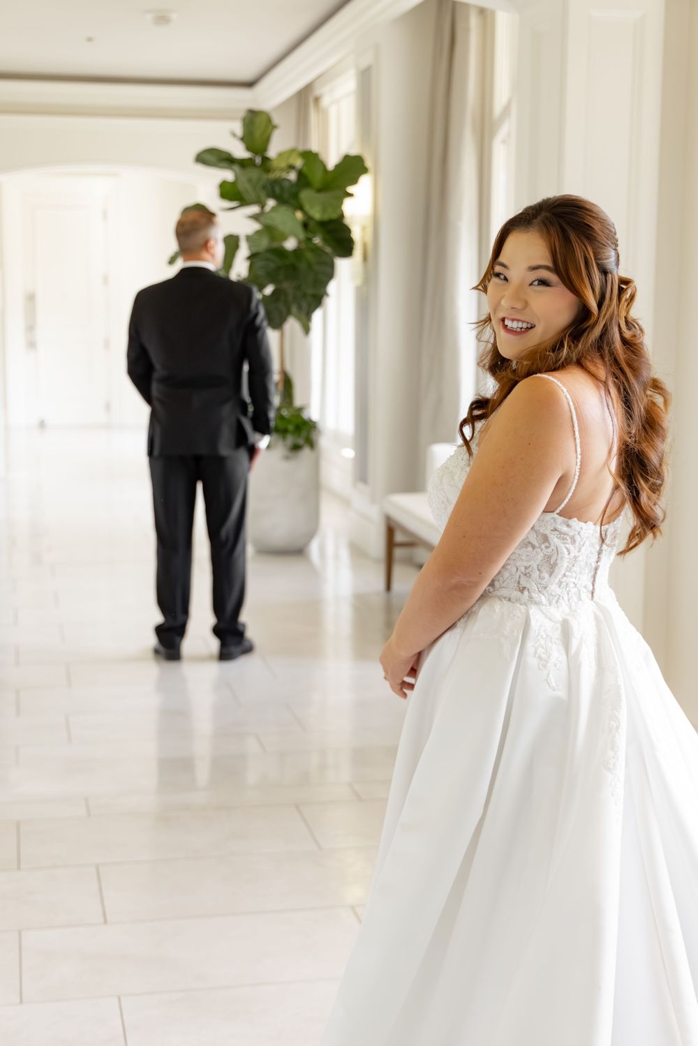 Smiling bride in white lace gown looks back while groom in tuxedo stands ahead in elegant hallway.