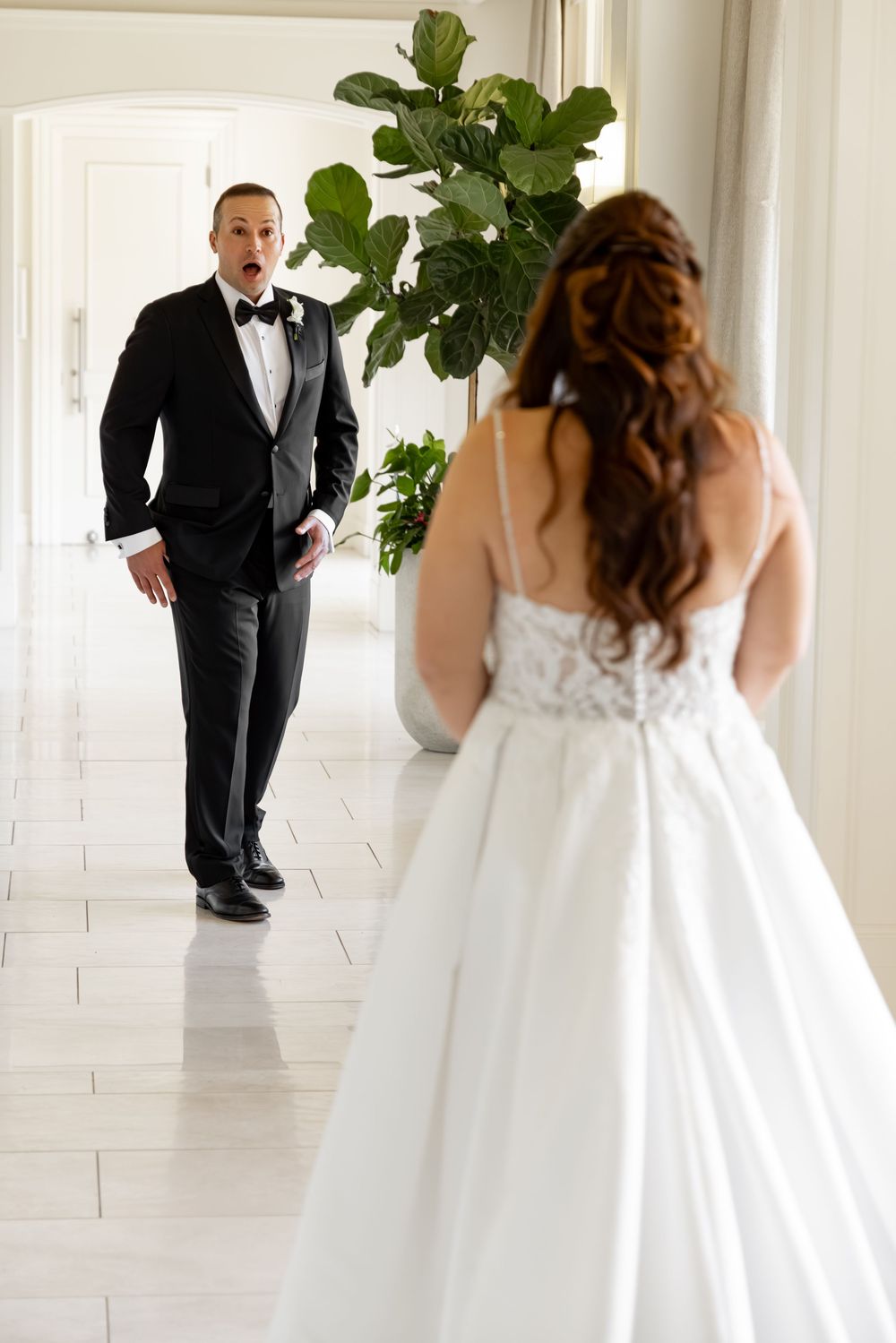 Groom in black tuxedo reacts with awe seeing bride in white lace wedding gown during first look moment.
