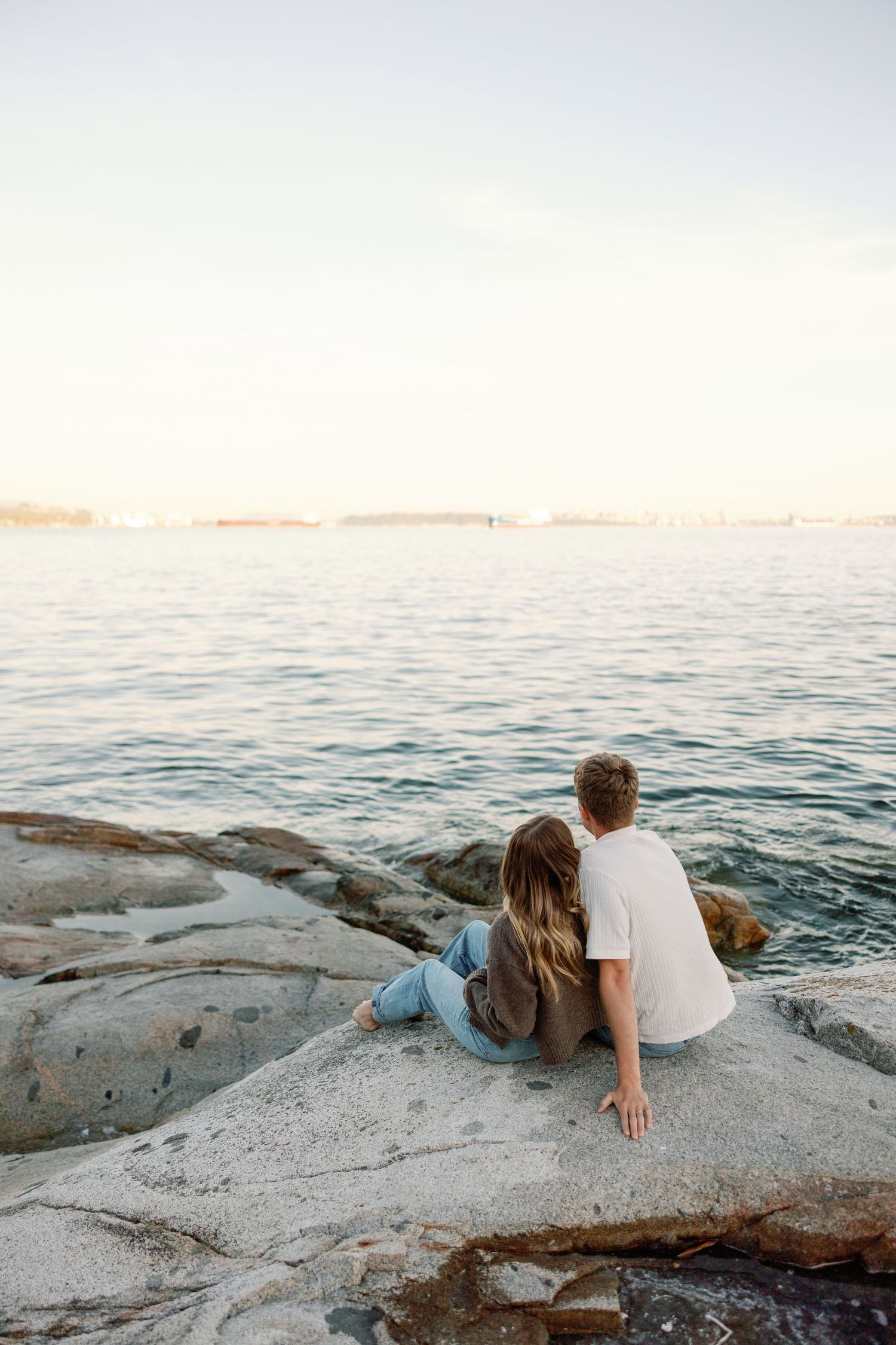 Couple sitting together on rocky shoreline gazing at calm ocean water during golden hour sunset.
