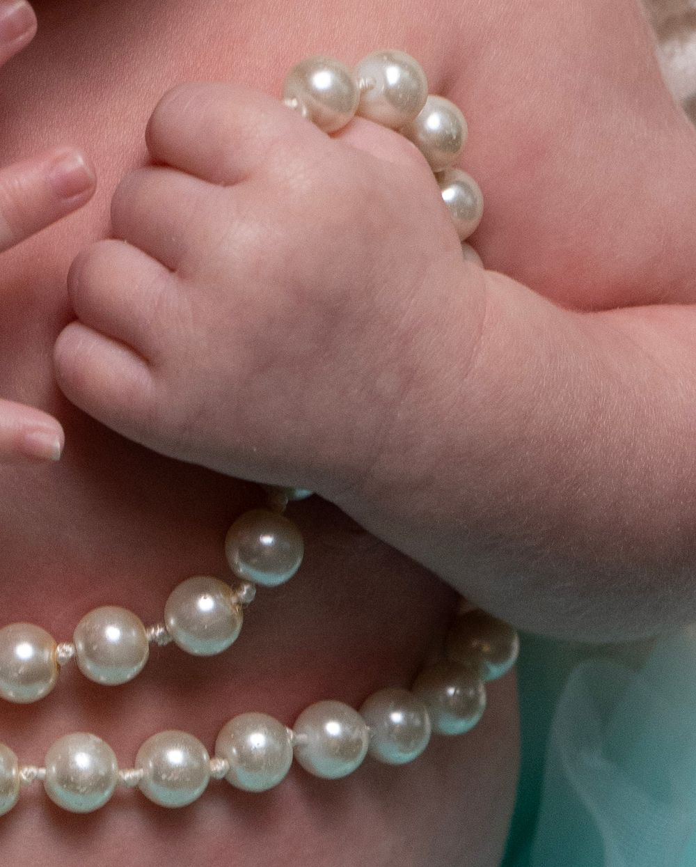 Close-up of a baby's tiny hand grasping a strand of white pearl necklace beads.