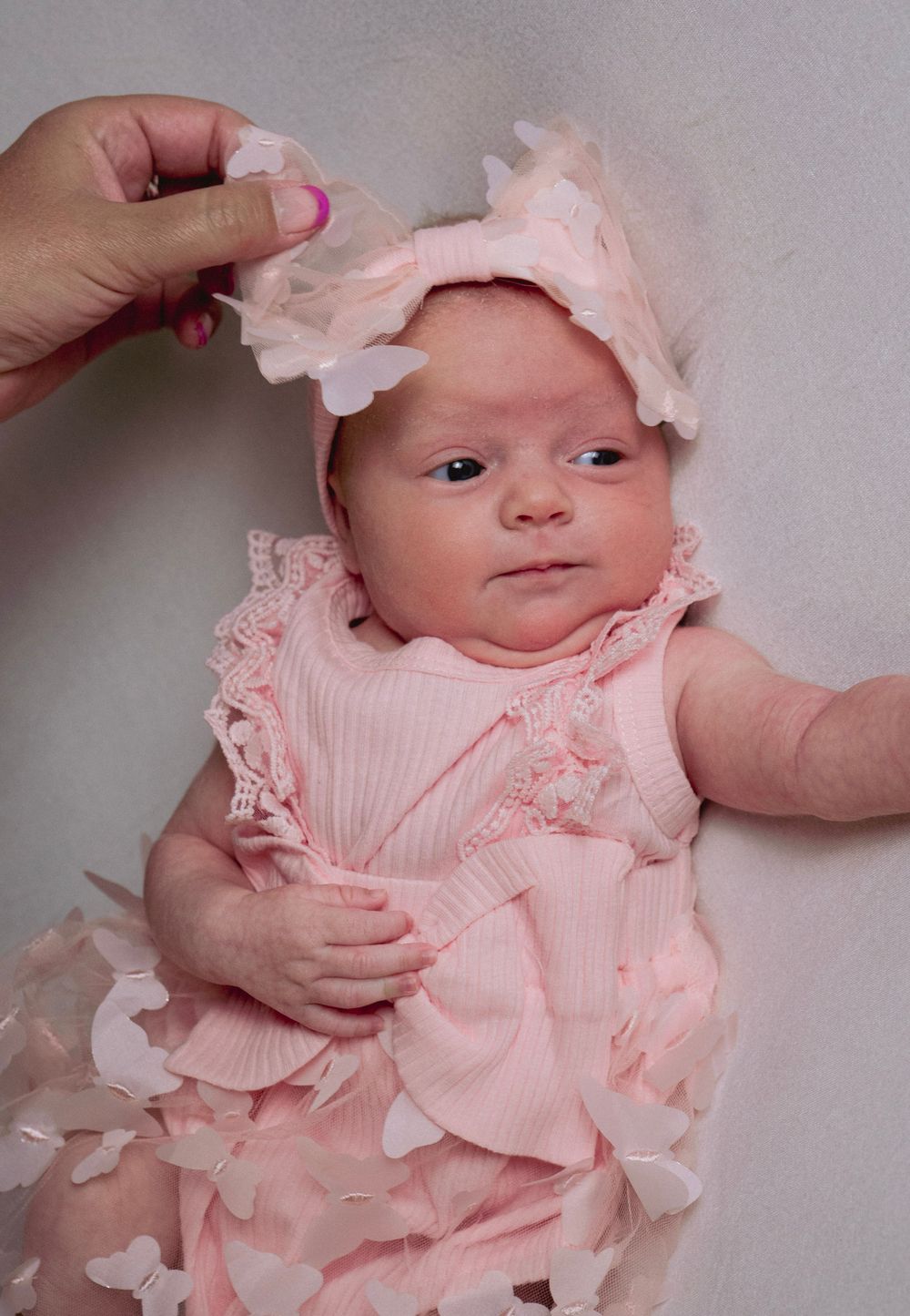 Newborn baby girl in pink ruffled outfit with butterfly headband, lying on white background.