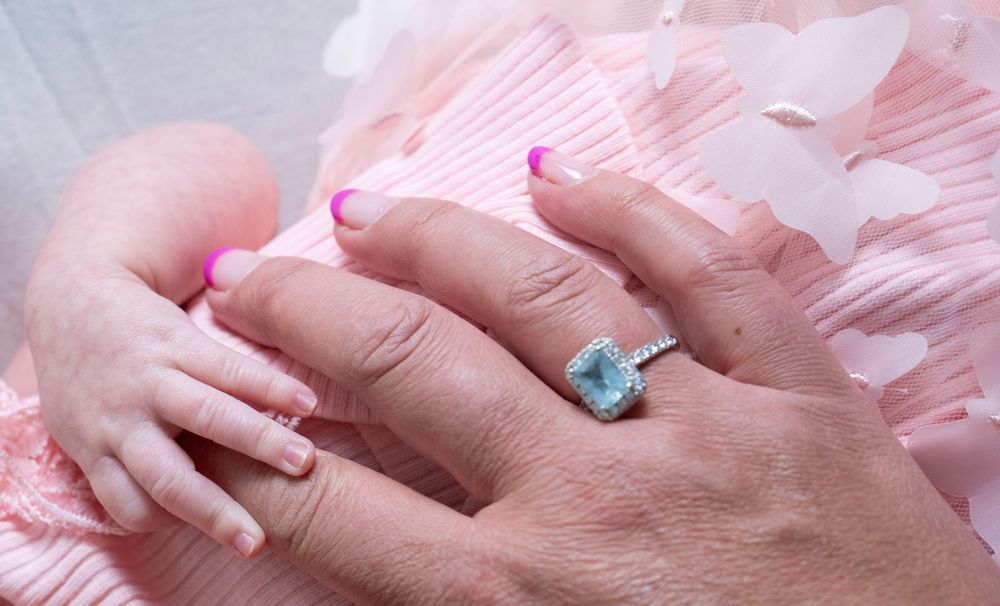 Adult hand wearing a blue gemstone halo ring rests next to a newborn baby's tiny hand on pink fabric.