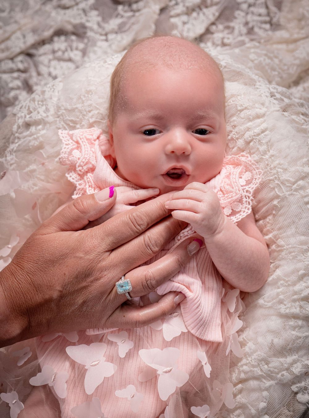Newborn baby girl in pink lace dress held by adult hands with pink nails and turquoise ring on white lace blanket.