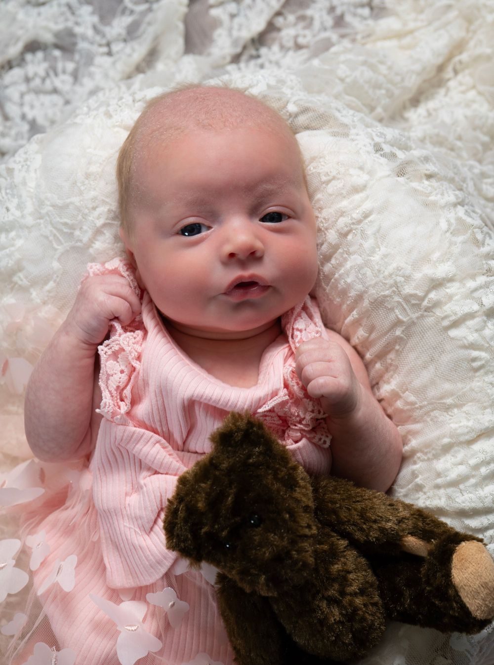 Newborn baby in pink dress lying on white lace blanket with a small brown teddy bear beside her.