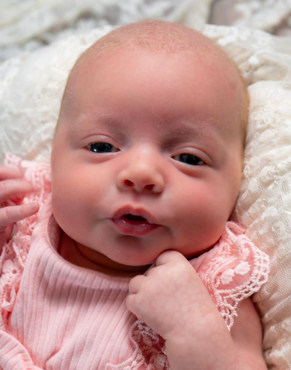 Adorable newborn baby girl in pink lace outfit lying on white blanket, with bright eyes and tiny fist near chin.