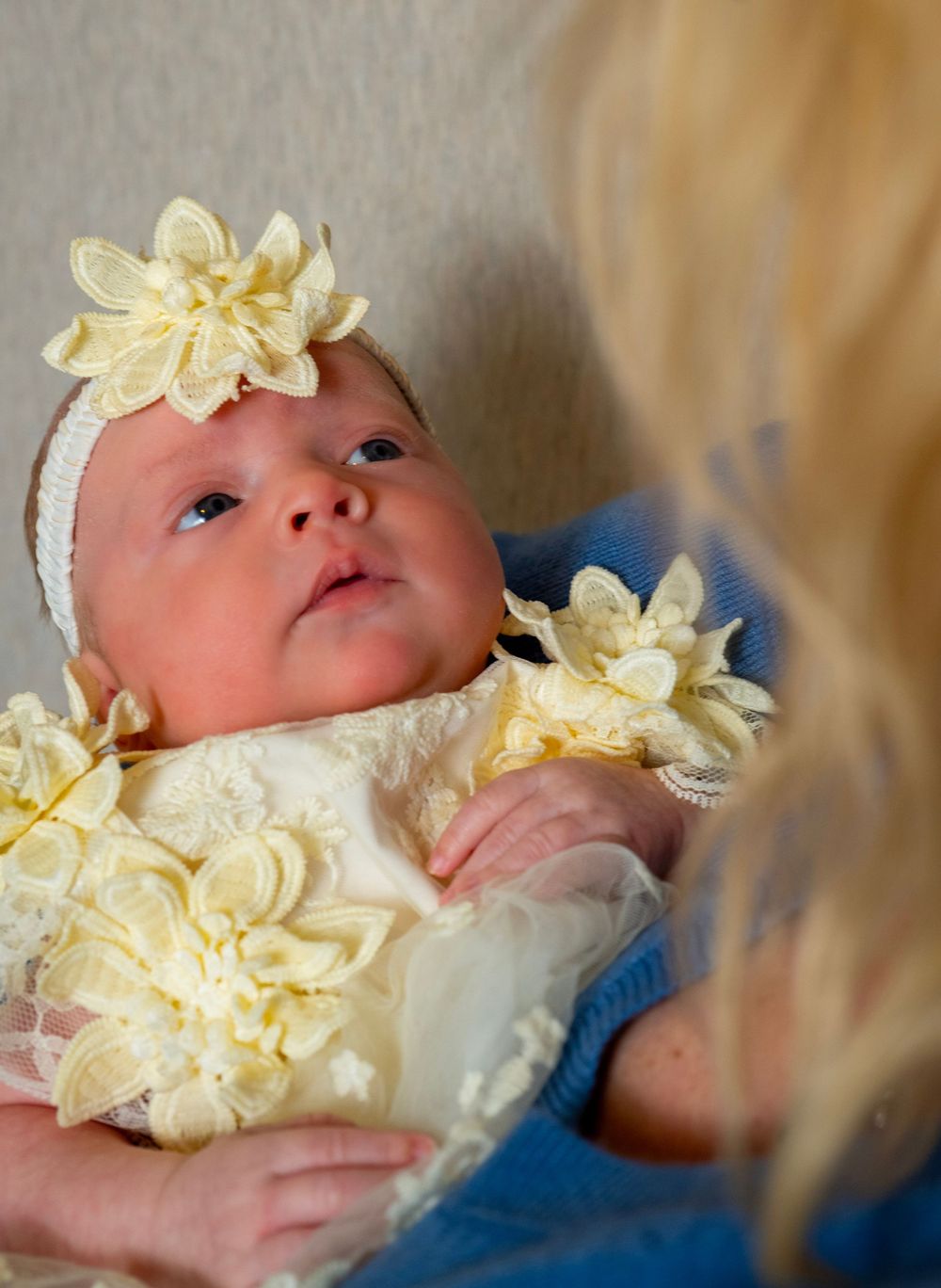 Newborn baby girl in white lace outfit with yellow flower headband being held by blonde woman.