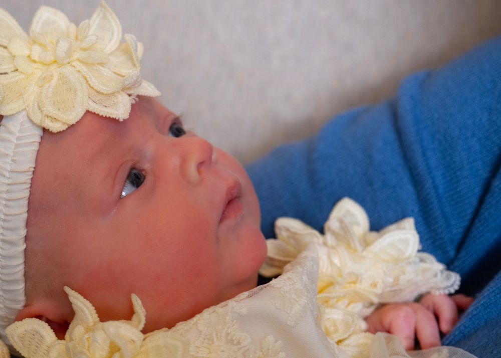 Newborn baby girl wearing a white knit hat with yellow flower, dressed in cream lace outfit.