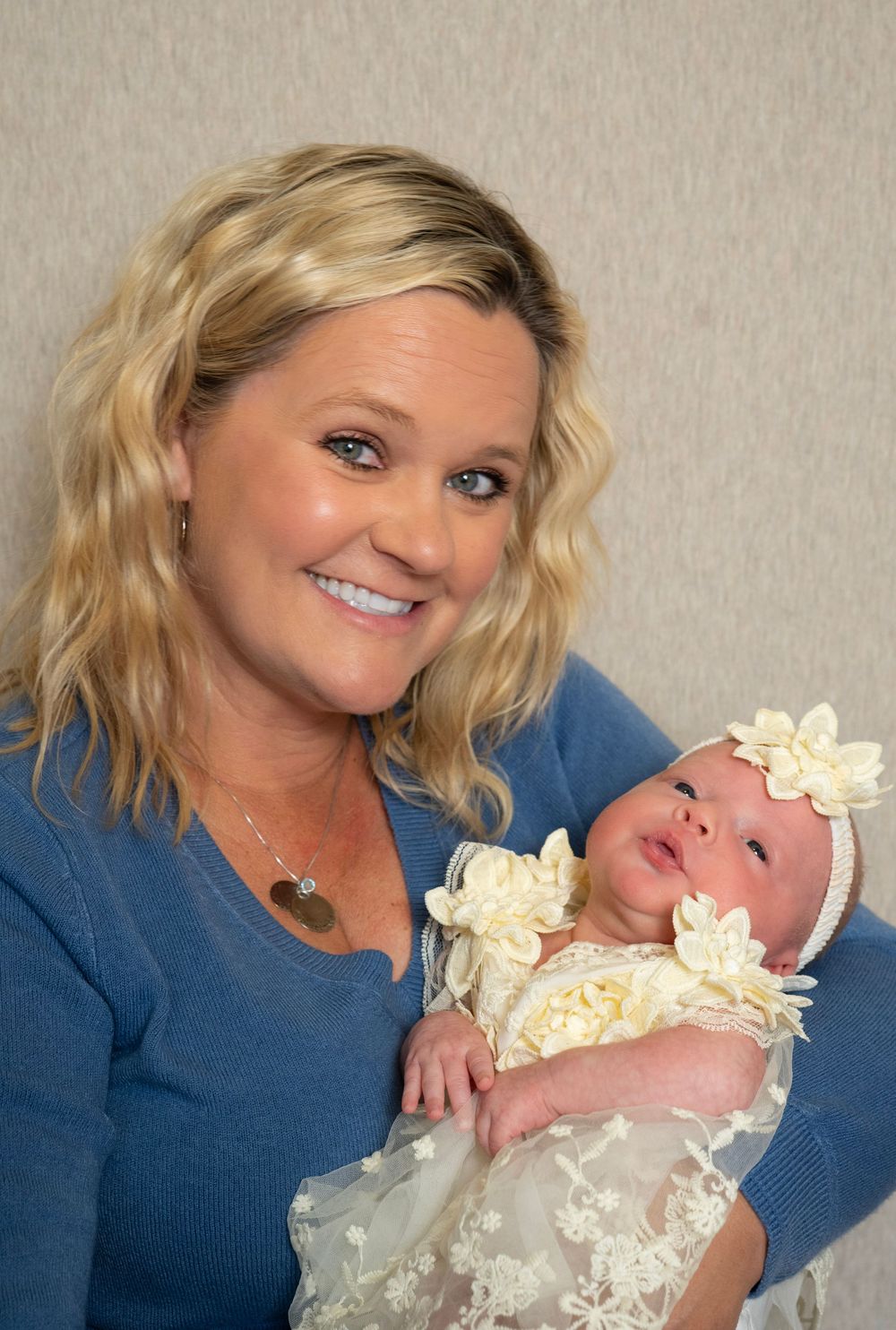 Smiling blonde woman in blue sweater holding a newborn baby girl in white floral dress and headband.