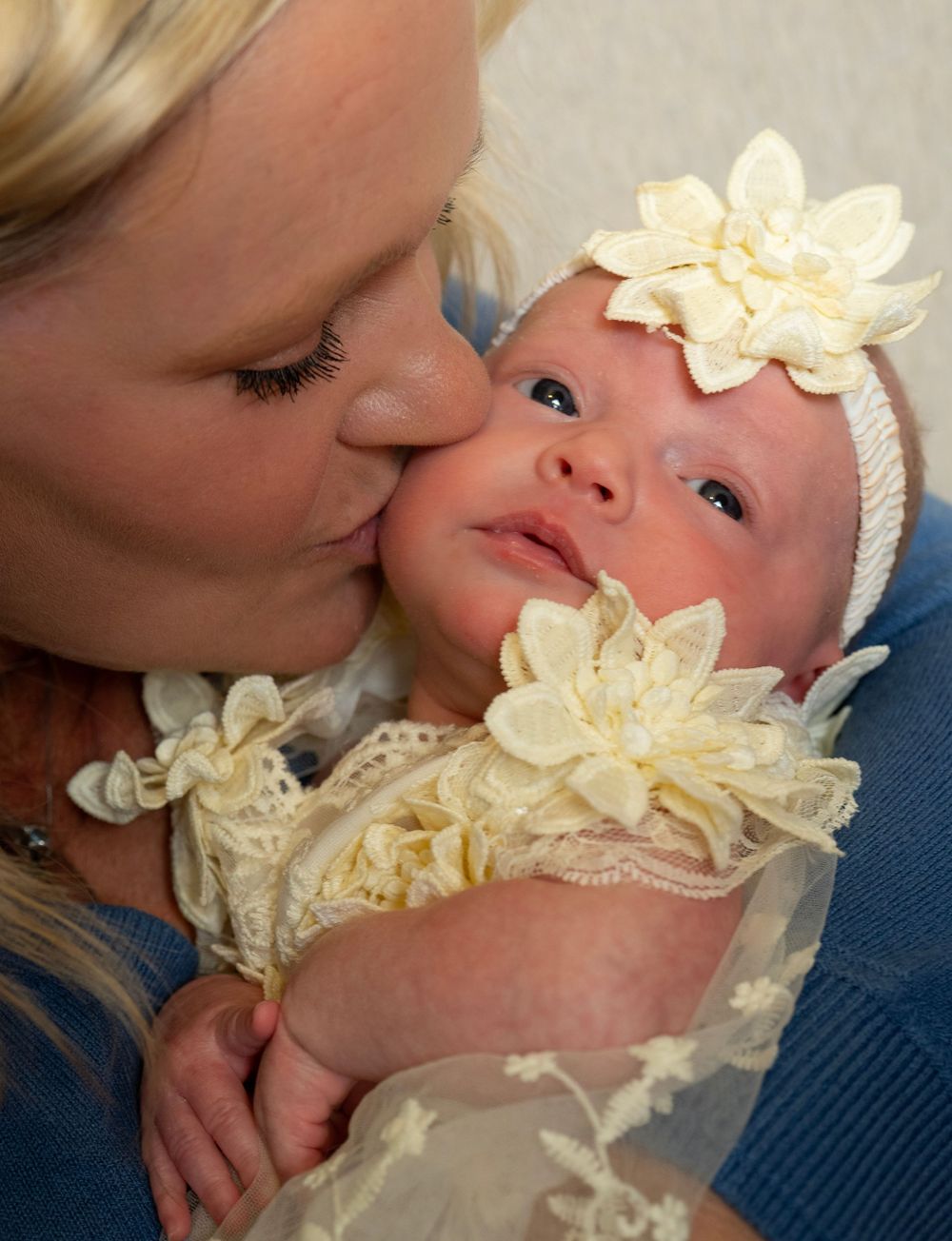 Mother kissing her newborn baby girl dressed in cream lace outfit with flower headband.