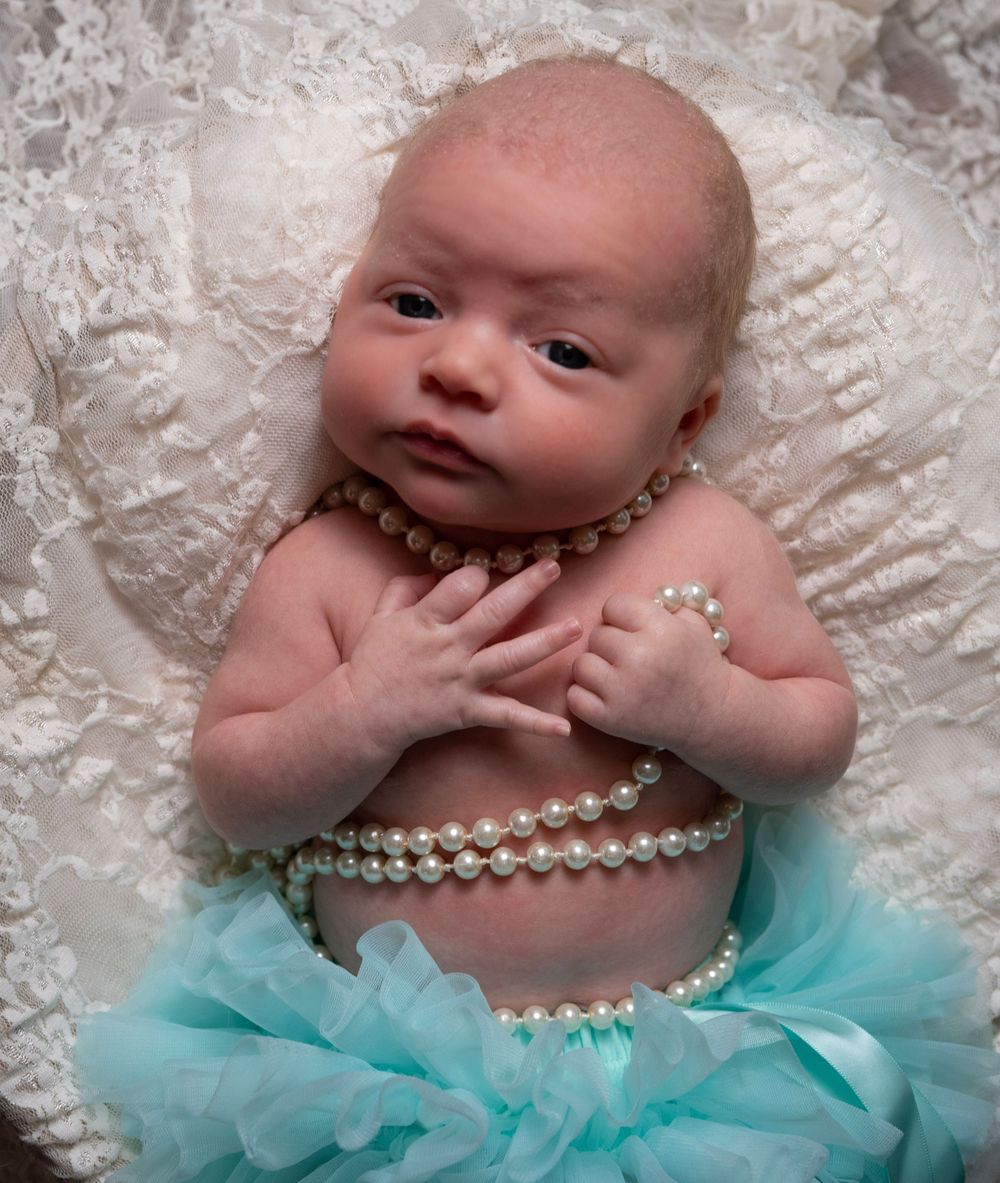 Newborn baby girl posed in teal tutu skirt with pearl necklace on white lace background.