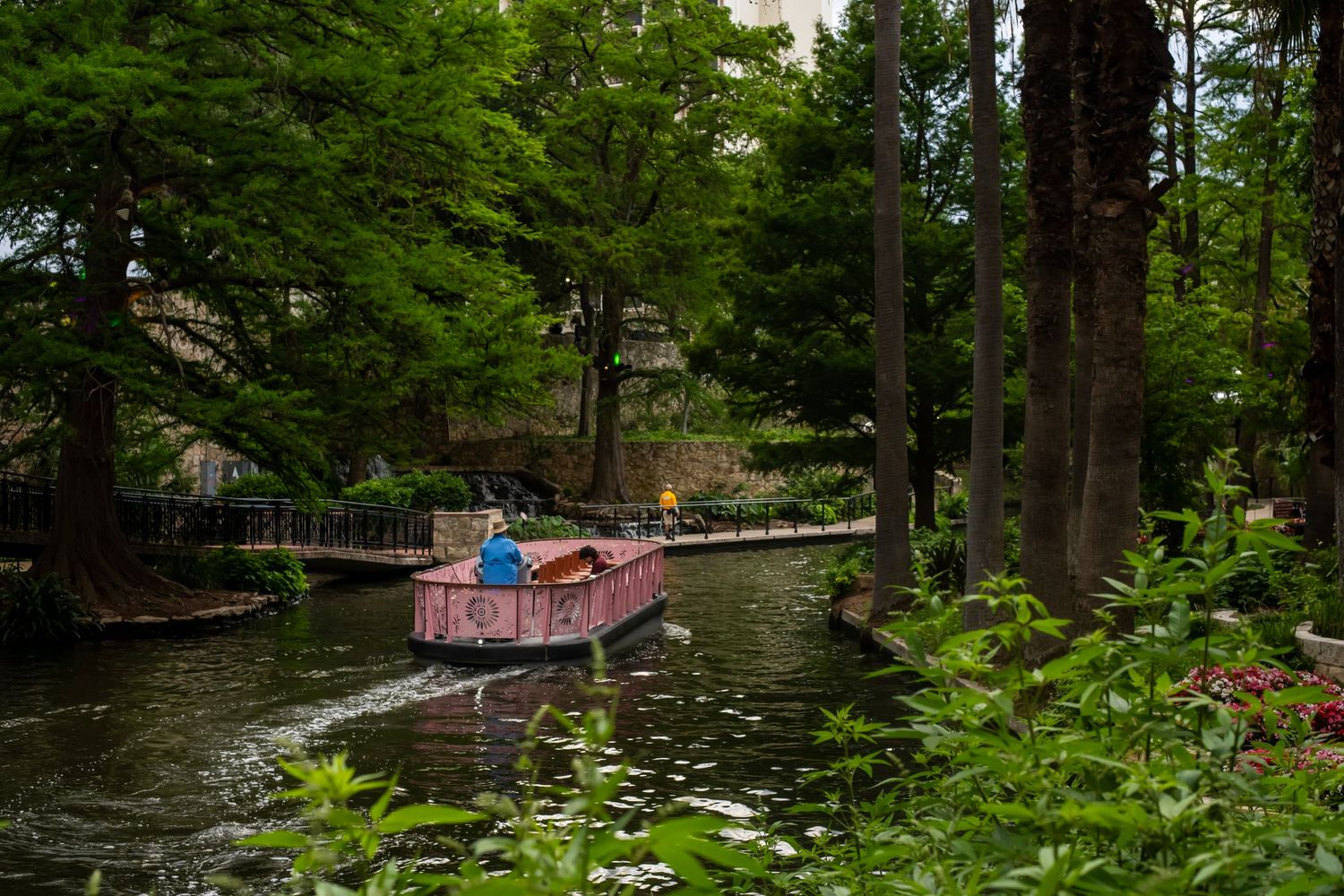 A pink boat glides through a lush green canal surrounded by tall trees in a scenic park waterway.