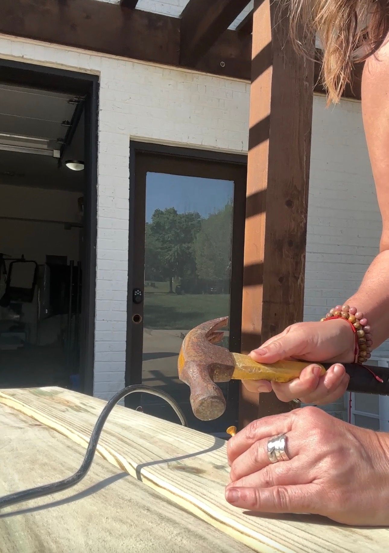 Woman using a hammer and nail on wooden boards outside a garage workshop, wearing a ring and red beaded bracelet.