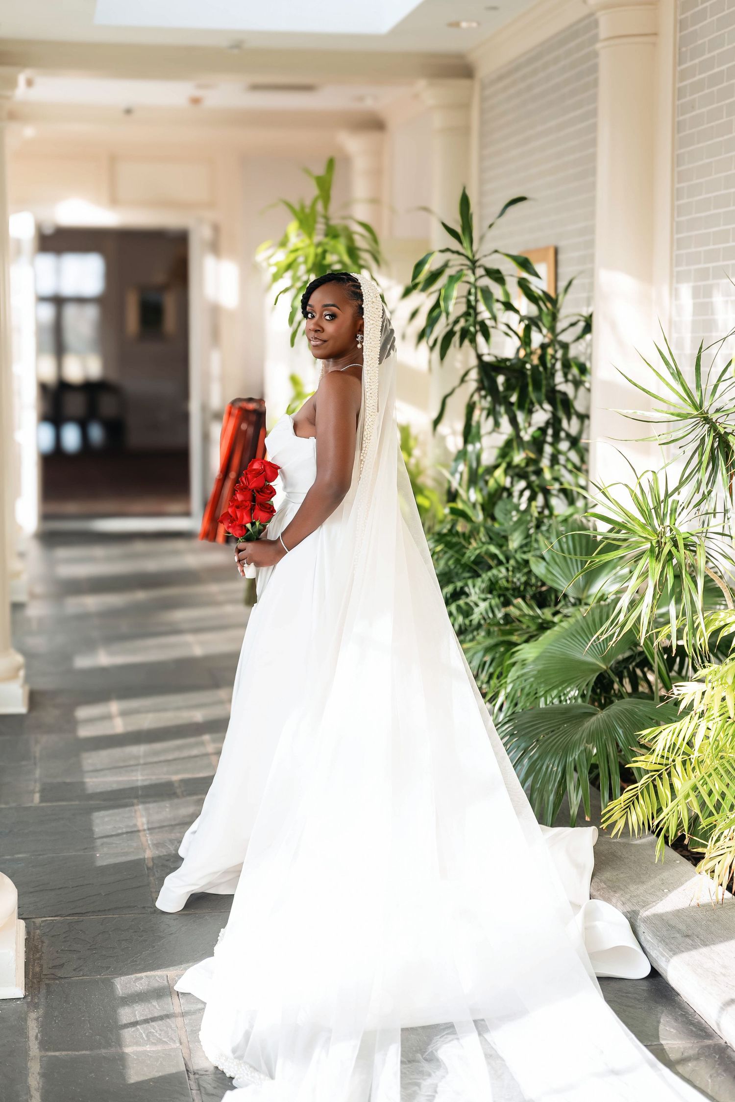 Black bride in white ballgown holding red roses stands in elegant venue corridor surrounded by tropical plants Deerfield Golf Club Newark De