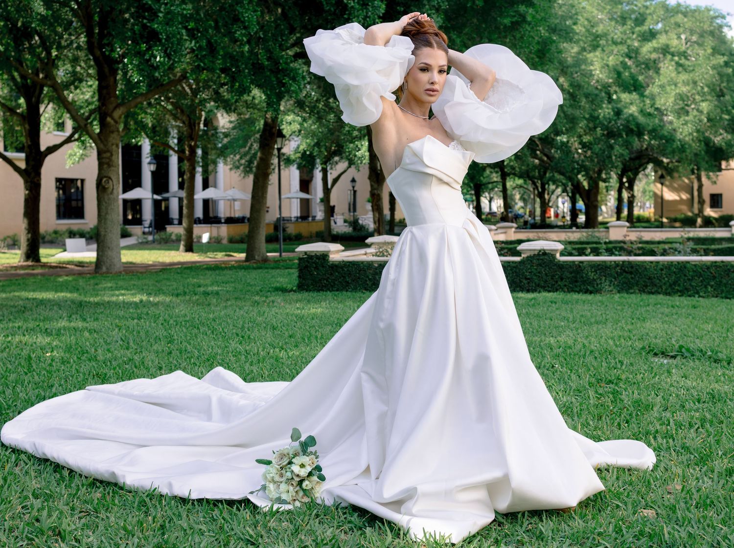 Bride in elegant white strapless ball gown with dramatic organza sleeves poses on lush green lawn outdoors.