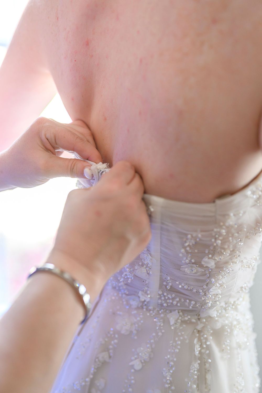 Helper fastening buttons on back of beaded white wedding dress on bride during bridal preparation.