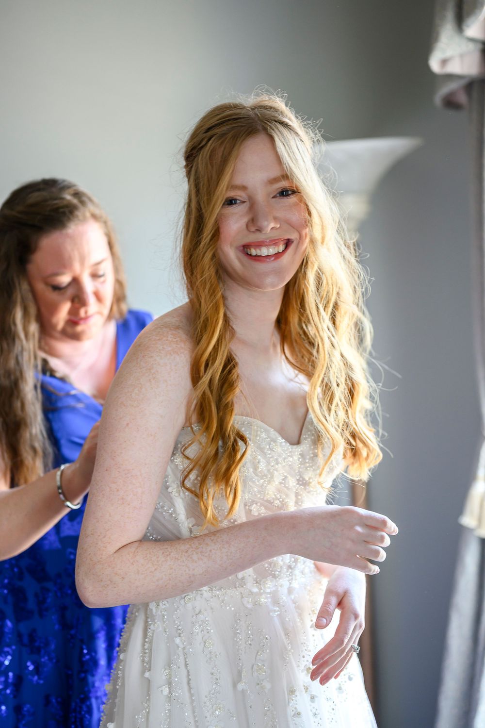 Smiling redhead bride in white gown being helped into dress by woman in blue sequin top.