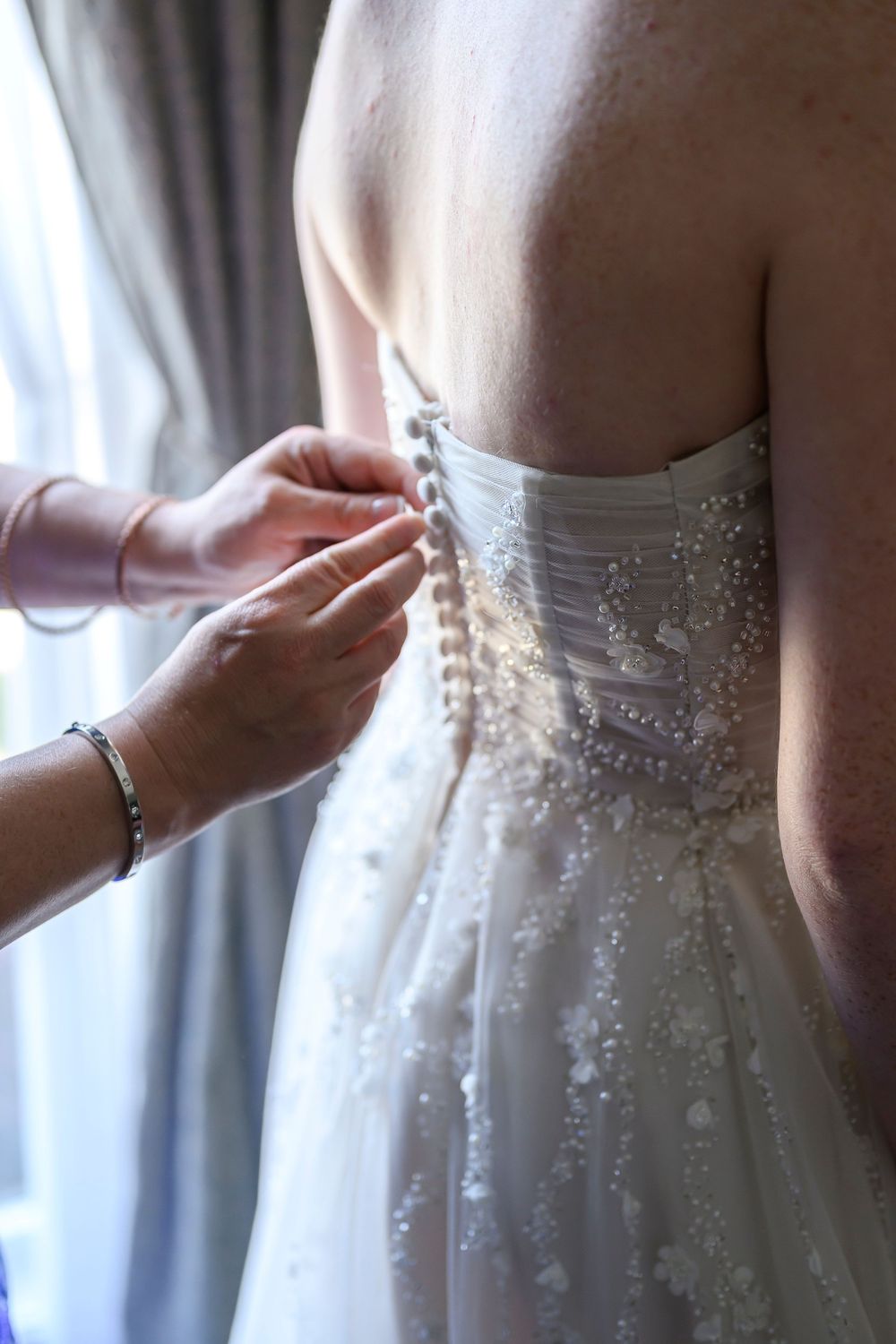 Hands fastening pearl buttons on the back of a beaded strapless white wedding dress near a window.