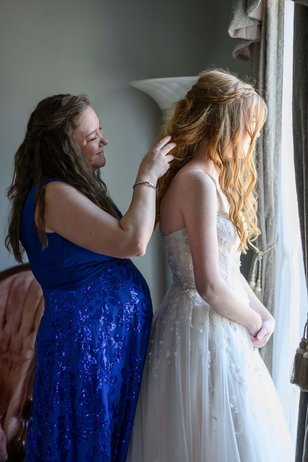 Woman in blue sequin dress helping bride in white gown with hair near window before wedding ceremony.