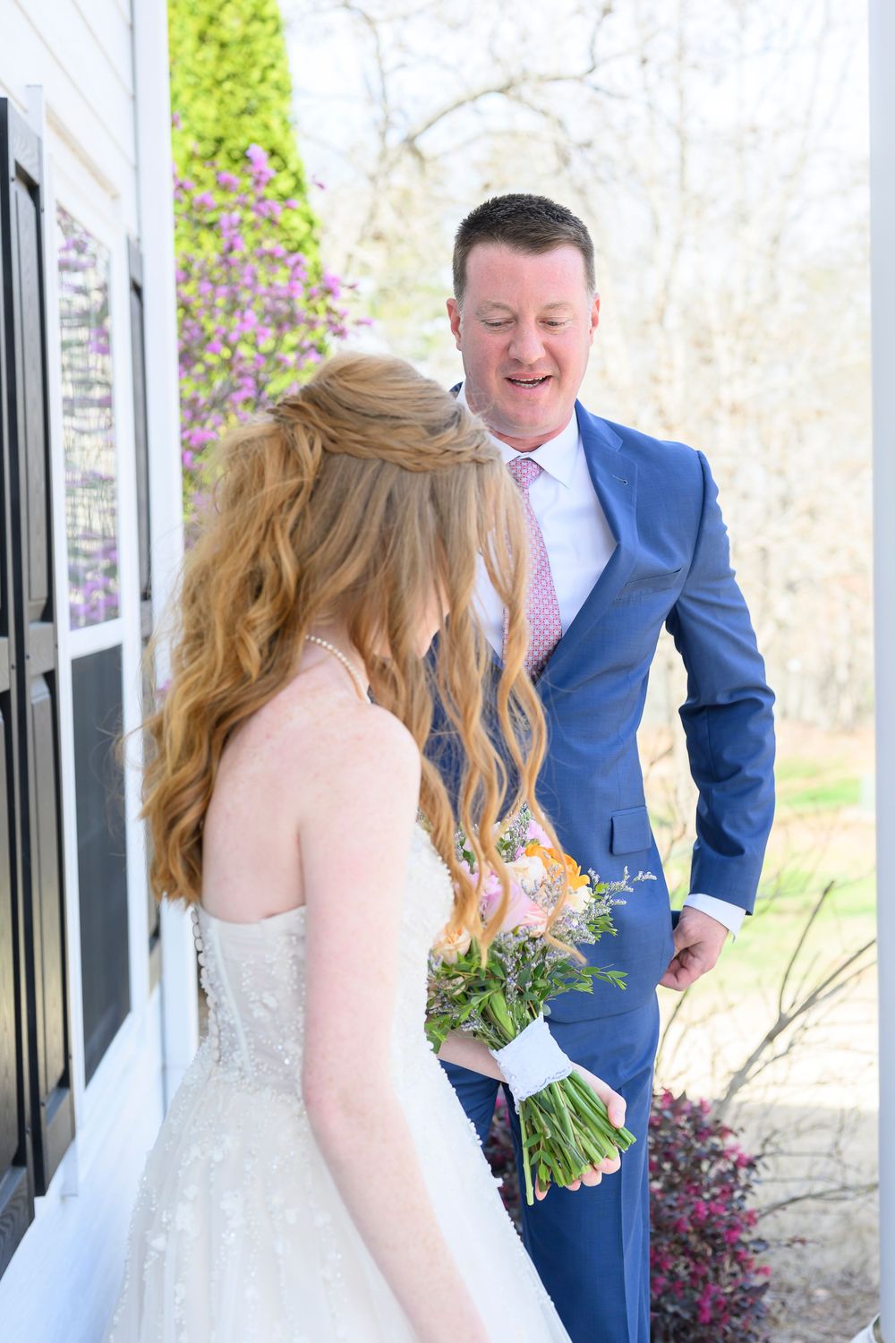 Groom in blue suit reacts with joy seeing bride in white gown holding colorful bouquet during wedding first look.