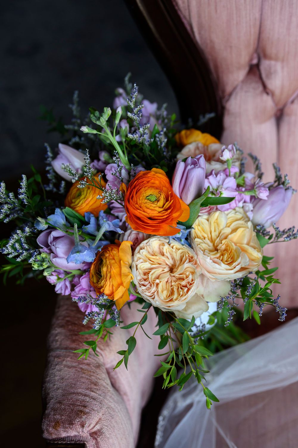 Colorful wedding bouquet with orange ranunculus, yellow roses, purple tulips, and blue flowers held against a pink velvet chair.