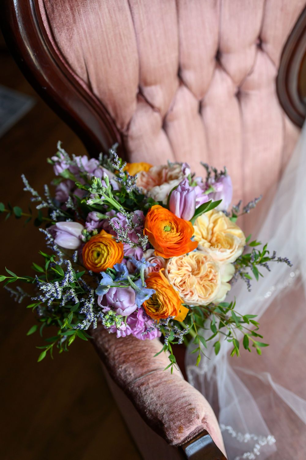 Colorful bridal bouquet with orange ranunculus, yellow roses, purple tulips, and greenery resting on a pink tufted chair.