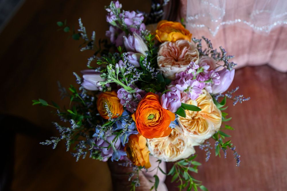 Vibrant bridal bouquet featuring orange ranunculus, peach roses, purple flowers, and green foliage held by a person.