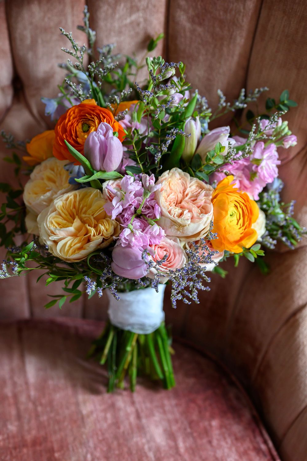 Colorful wedding bouquet with orange ranunculus, yellow roses, pink tulips, and wildflowers resting on a pink velvet chair.