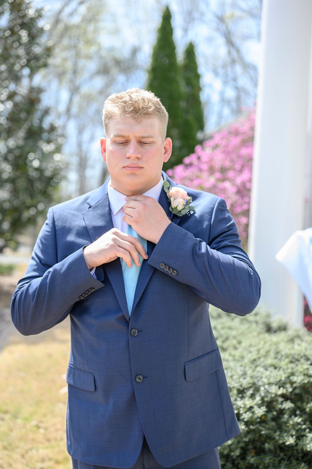 Young man in navy blue suit adjusting light blue tie, wearing boutonniere, outdoors at a wedding ceremony.