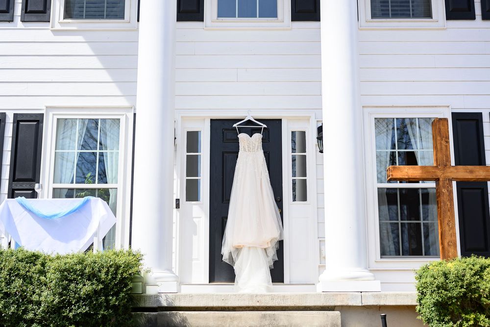 Wedding dress hanging on hanger outside white colonial home with black shutters and front door.