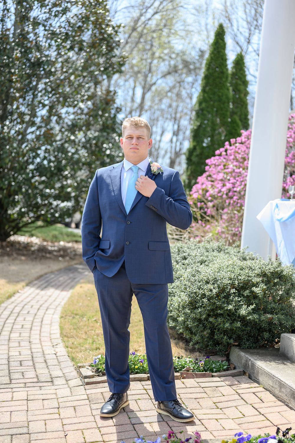 Groom in navy blue suit with light blue tie and boutonniere poses on brick path surrounded by spring garden blooms.